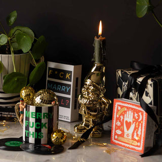 A table of gifts with a box of matches, gold gnome candle holder, Sweary stripe Christmas mug, card game and a wrapped present and card in the background