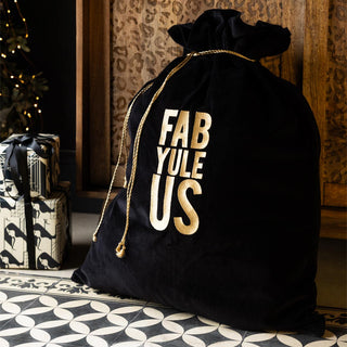 The Luxury Yule Us Velvet Christmas Sack displayed on the floor with some presents in front of a leopard print sideboard and some fairy lights.