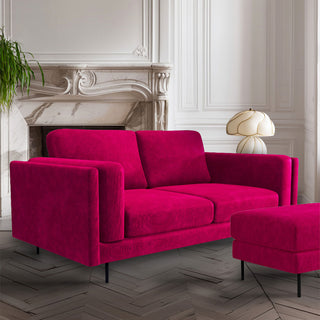 A bright pink sofa styled in a living room with the matching footstool, with a lamp and plant in front of a fireplace.