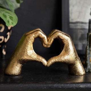 The Antique Gold Heart Hands Ornament displayed on a black surface with a plant.