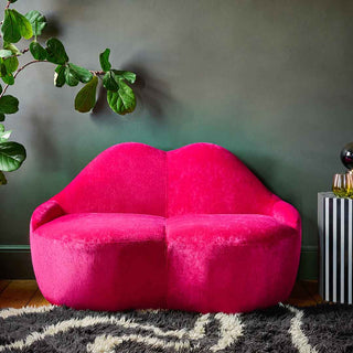 A striped sofa in the shape of some lips, styled on a rug in front of a dark wall with a cushion and a table lamp on a table.