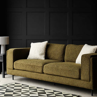 The Olive Chunky Boucle Sofa styled with two cream cushions, a lamp, side table and geometric rug, in front of a black panelled wall. 