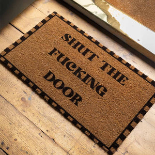 The Shut The Fucking Door Doormat displayed on a wooden floor by an open door.