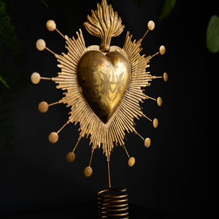 The Milagro Heart Christmas Tree Topper displayed on a dark surface with greenery in the background.