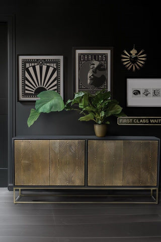 Gold and matt black cabinet with metal frame styled on a dark wood floor in front of black and white prints, a Milagro heart and a leafy green plant