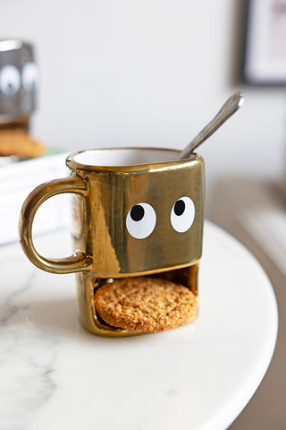 Gold cookie holding face mug on a white side table. The mug has a silver spoon in it and the cookie compartment features a oat biscuit. The novelty mug has two eyes.