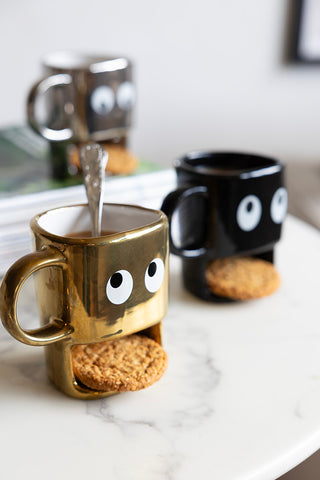 Three cookie holding face mugs in gold, black and silver. The gold face mug is at the forefront with the black novelty mug in the background and the silver face mug slightly blurred in the left hand corner.