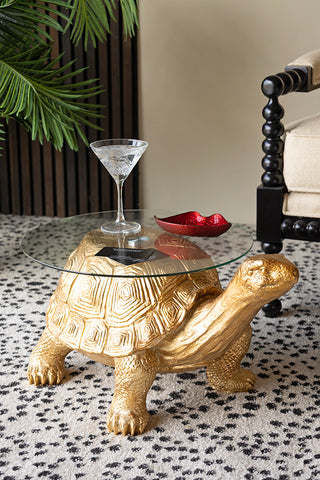 The Gold Tortoise Side Table styled with a glass and trinket dish on, next to a chair and a plant.