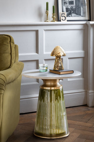 The Green Tapered Glass & Marble Side Table with some accessories on, styled next to a sofa, with some artwork and candlesticks on a shelf in the background.