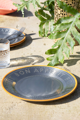 The Metal & Orange Rim Bon Appetit Dinner Plate styled on an outdoor table with a plant and various other dining accessories.