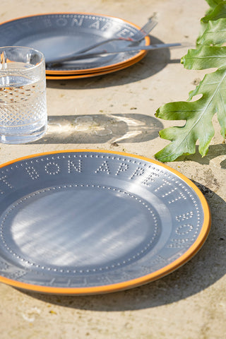 The Metal & Orange Rim Bon Appetit Dinner Plate styled on a table, with more plates in the background, with a plant, cutlery and glass.