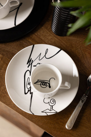 The Male Face Coffee Cup & Saucer styled on a table with other tableware and a plant.