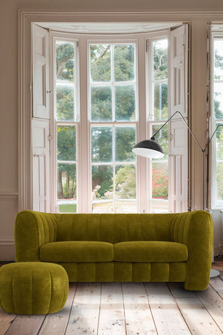 The Bowie Medium Sofa In Luxe Kneedlecord Velvet Vintage Green styled in front of a large window with a matching pouffe in the front