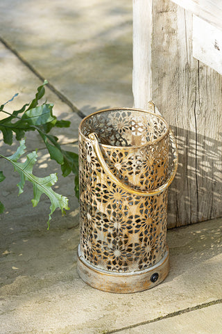 The Rustic-Style Outdoor Lantern styled on a patio floor next to a plant and some wooden furniture.