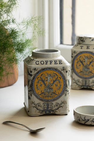 The Coffee Storage Jar from the Set of 2 Ceramic Tea & Coffee Storage Jars, with the lid off, styled with a spoon, plant, wooden board and the Tea version in the background.