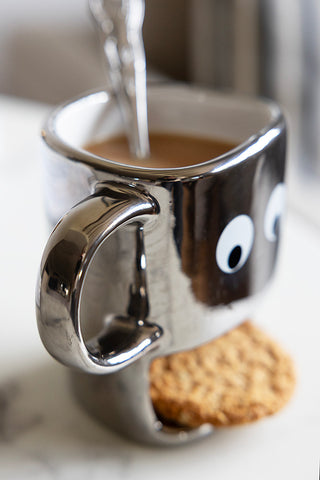 Side view of the silver mug. The mug is filled with tea with a silver teaspoon inside. On the outside of the novelty mug is big white googly eyes and a slot where the mouth is meant to be, with a gap big enough to store a biscuit.