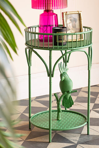 The Tropical Green Parrot Side Table styled with a lamp, photo frame and candle on, with a plant to the side of the shot.