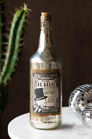 The Vintage-Style Poison Ornamental Glass Bottle on a table with some disco balls and a cactus in the background.