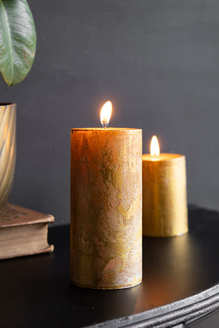 The Shiny Gold Pillar Candle - Tall displayed with the smaller version on a black sideboard, with a book and a plant.