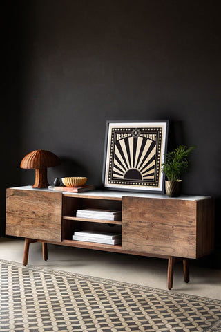 The Acacia Wood & Marble TV Unit styled with a lamp, art print and various accessories on, in front of a dark wall.