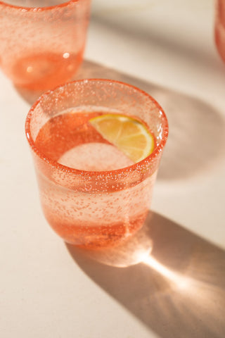 A lifestyle image of a coral pink picnic tumbler glass with water and a lemon slice in it. 