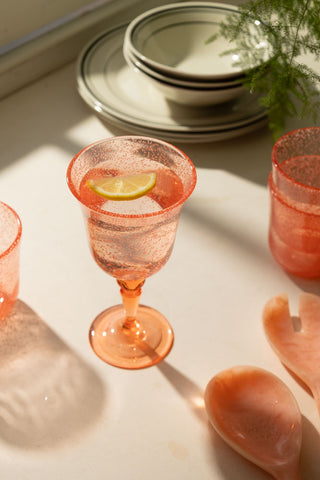 A lifestyle image of an acrylic coral pink wine glass with water and a lemon in it. 