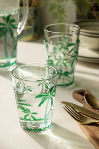 A lifestyle image of an acrylic green palm tree tumbler glass next to a chopping board with gold cutlery on it. 