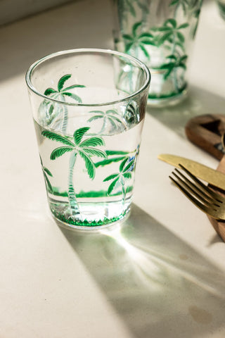 A lifestyle image of a clear acrylic tumbler glass with green palm trees on it. 