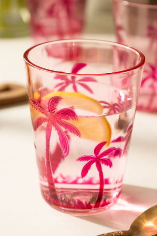 A lifestyle image of an acrylic pink palm tree tumbler glass with water and a slice of lemon in it. 