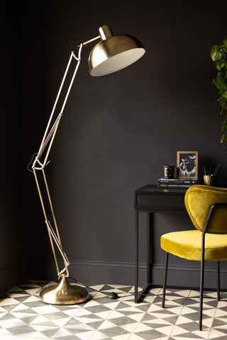 The Adjustable Gold Task Floor Lamp displayed in a dark room with a desk, chair, plant and various accessories.