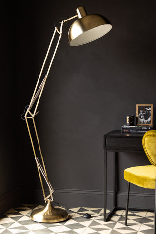 The Adjustable Gold Task Floor Lamp displayed in a dark room next to a desk and chair with various accessories.