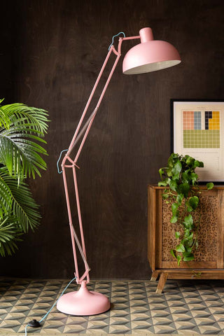 The Adjustable Pink Task Floor Lamp with Blue Flex displayed in front of a dark wall, next to a sideboard, art print and some plants.