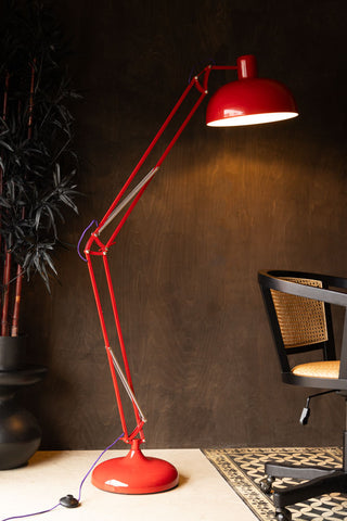 The Adjustable Red Task Floor Lamp with Purple Flex switched on, displayed in front of a dark wall next to a wicker desk chair and a large plant. 