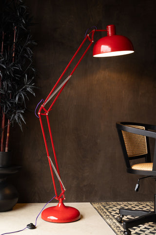 The Adjustable Red Task Floor Lamp with Purple Flex switched on and styled in front of a dark wall, next to a large plant and a desk chair.