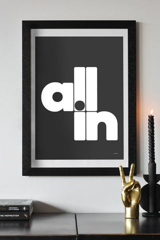 The All In Black & White Art Print - Unframed, styled in a frame on a white wall, above a black surface styled with books, a gold hand ornament and a lit candle in an abstract candle holder.