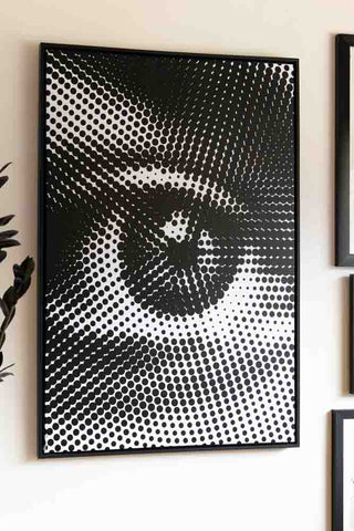 A canvas with an eye design on a white wall, next to other art and a plant.