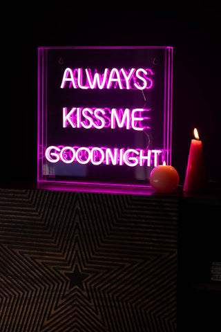 The Always Kiss Me Goodnight Neon Light Box styled switched on in a dark room, next to some pink candles.