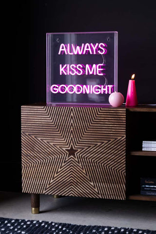 The Always Kiss Me Goodnight Neon Light Box illuminated, styled on a wooden unit next to two lit pink candles.