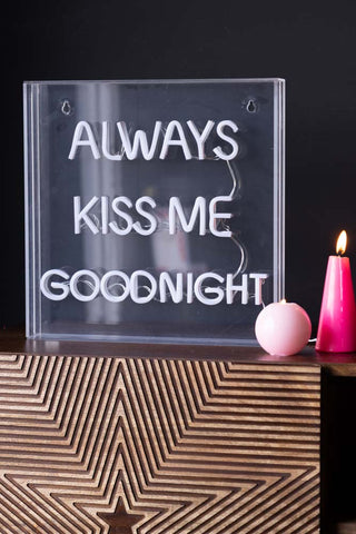 The Always Kiss Me Goodnight Neon Light Box switched off, styled on a wooden unit with two lit pink candles.