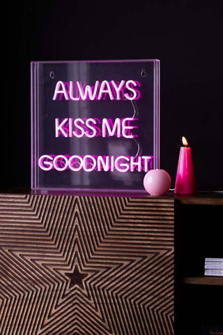 The Always Kiss Me Goodnight Neon Light Box switched on and styled on a wooden unit with some pink candles.