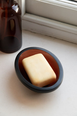 The Apothecary-Style Amber Glass Soap Dish styled with a bar of soap inside, with a matching amber glass soap dispenser in the background.