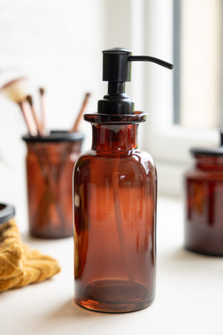 The Apothecary-Style Amber Glass Soap Dispenser - 300ml on a window ledge with matching amber glassware items in the background.