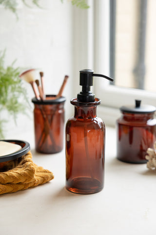 The Apothecary-Style Amber Glass Soap Dispenser - 300ml displayed on a window ledge with matching amber glassware items, with a plant and other bathroom accessories.