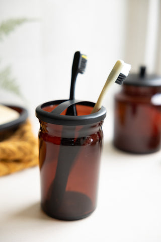 The Apothecary-Style Amber Glass Toothbrush Holder displayed with toothbrushes inside and other bathroom accessories in the background.