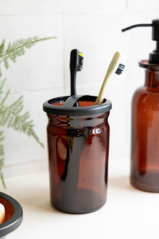 The Apothecary-Style Amber Glass Toothbrush Holder with toothbrushes inside, styled with matching amber glassware and a plant.