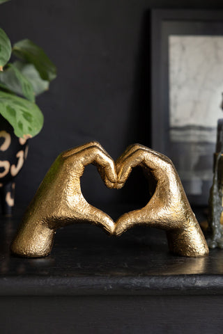 The Antique Gold Heart Hands Ornament displayed on a black sideboard with a plants and some other home accessories.