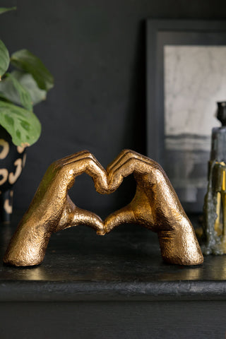 The Antique Gold Heart Hands Ornament styled on a black surface with a plant, photo frame and some candlestick holders in the background.