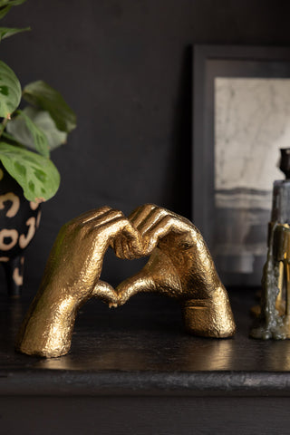 The Antique Gold Heart Hands Ornament seen from an angle, styled on a black surface with a plant and some home accessories.