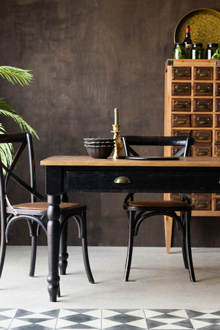 The Apothecary Dining Table styled with tableware, chairs, a plant and a tall storage unit.