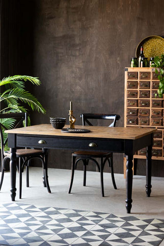 The Apothecary Dining Table styled with various furniture and home accessories, in front of a dark wooden wall with plants.